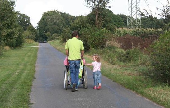 In Rückansicht: Eine Person schiebt einen Rollstuhl und nebenher läuft ein kleines Kind.