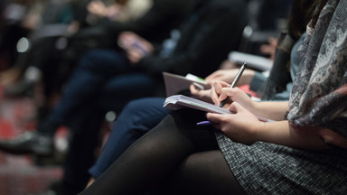 Der Bildausschnitt zeigt einen Teil eines Publikums auf einer Konferenz, die sich Notizen machen.