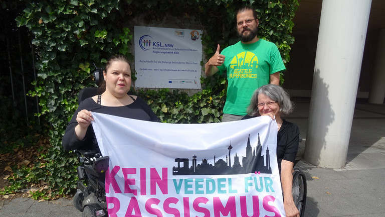 Drei KSL-MitarbeiterInnen präsentieren die Fahne von "Kein Veedel für Rassismus" vor dem KSL Köln.