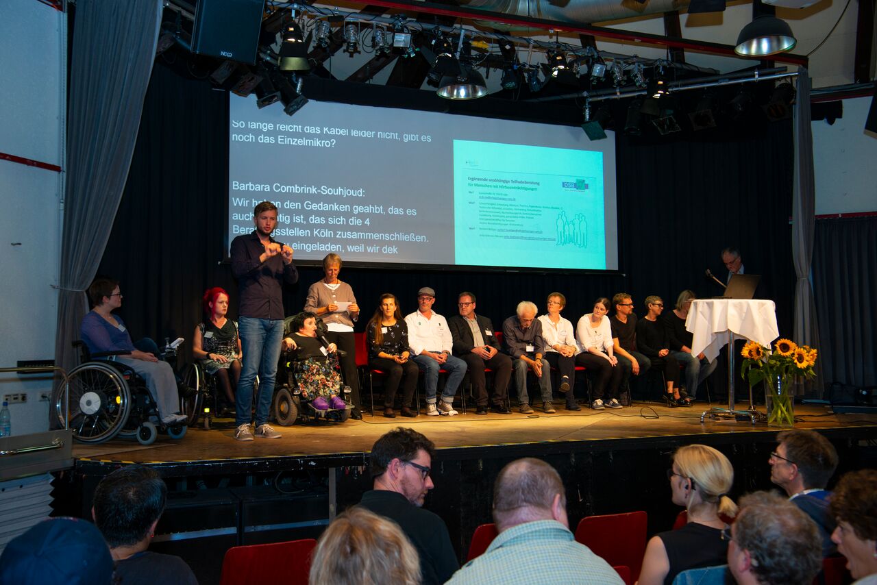 Mehrere Beraterinnen und Berater auf der Bühne mit Moderatorin und Gebärdensprachdolmetscher - im Hintergrund Power-Point-Präsentation.r. © Martin Lässig