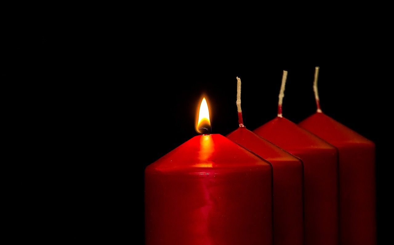 Das Bild zeigt vier rote Kerzen, von denen eine brennt.