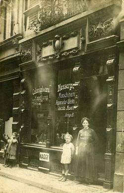 Ein altes Foto aus den 20er-Jahren. Es zeigt das Geschäft von Jacob Auen. Eine Frau und ein Kind stehen im Eingang und schauen in die Kamera. Zwei weitere Kinder stehen vor dem Schaufenster des Geschäfts.