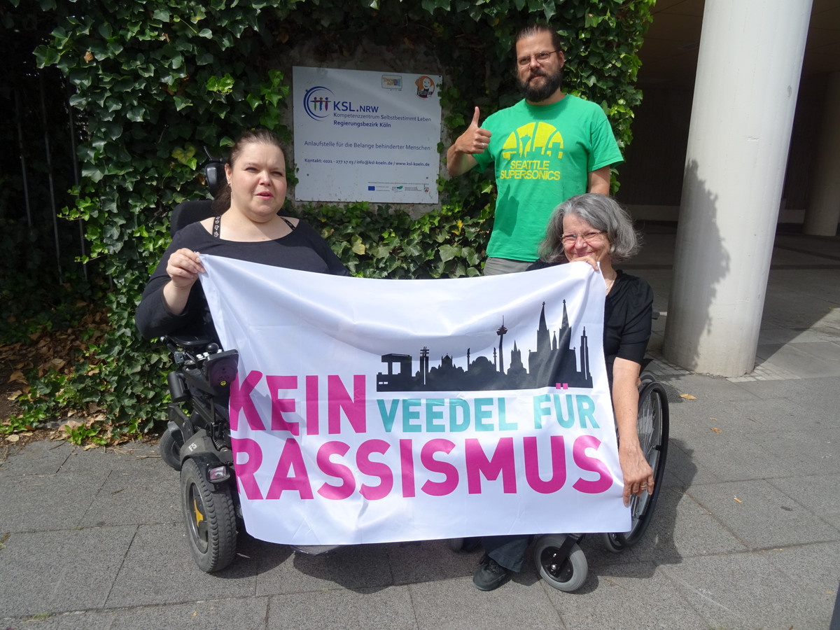 Drei KSL-MitarbeiterInnen präsentieren die Fahne von "Kein Veedel für Rassismus" vor dem KSL Köln.