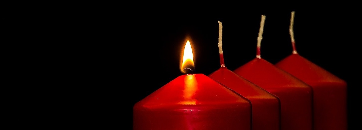 Das Bild zeigt vier rote Kerzen, von denen eine brennt.