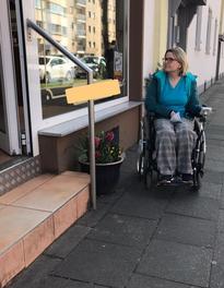 Eine Rollstuhlfahrerin steht vor eine Geschäft mit Stufe.