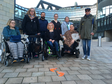 Mitarbeiter*innen von SL Behinderter Köln mit der Abgeordneten Woestmann vor dem Landtag NRW und dem Paar orangener Fußabdrücke