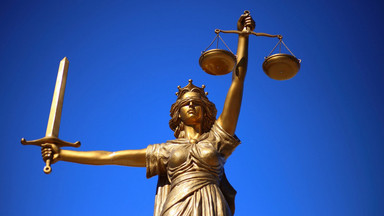 Statue der Justizia mit verbundenen Augen, Schwert in der rechten, Waage in der linken Hand vor blauem Himmel