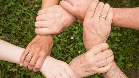 Mehrere Personen greifen mit nur einer Hand an das Handgelenk einer weiteren Person und stehen so, dass sie mit ihren Händen einen geschlossenen Kreis bilden.