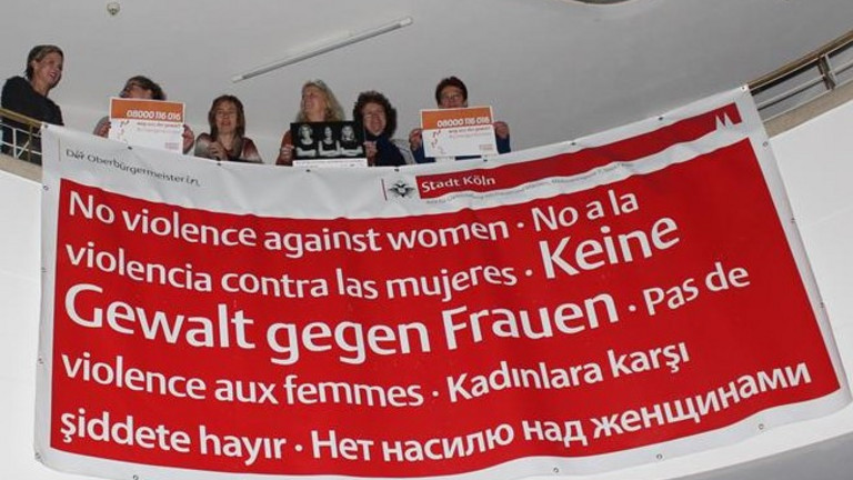 Das Bild zeigt Aktivistinnen auf einer Empore mit einem Bann "Keine Gewalt gegen Frauen" (in mehreren Sprachen).