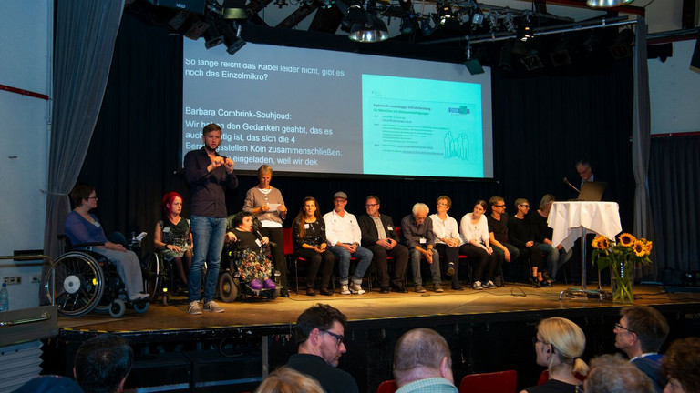 Mehrere Beraterinnen und Berater auf der Bühne mit Moderatorin und Gebärdensprachdolmetscher - im Hintergrund Power-Point-Präsentation.r. © Martin Lässig