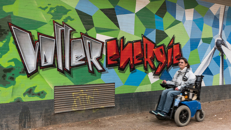Silke Katzfuß fährt in ihrem E-Rolli an einem Graffiti vorbei: "Voller Energie"