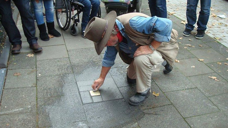 Ein Mann säubert einen frisch gesetzten Stolperstein auf dem Bürgersteig.