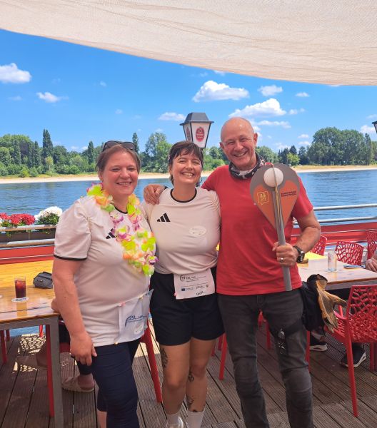 Ira Vogt und Lena Ingenhaag stehen Arm in Arm mit einem Vertreter von Biker for Kids Cologne e.V. Dieser hält den Staffelstab vor sich. Alle drei lächeln in die Kamera.