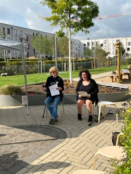 Jeanette Severin von Selbstbestimmt Leben e.V. und Mirjam Thomše, Behindertenbeauftragte der Stadt Köln, sitzen im Innenhof und halten Zettel in der Hand. Jeanette Severin hält ein Mikrophon in der Hand.