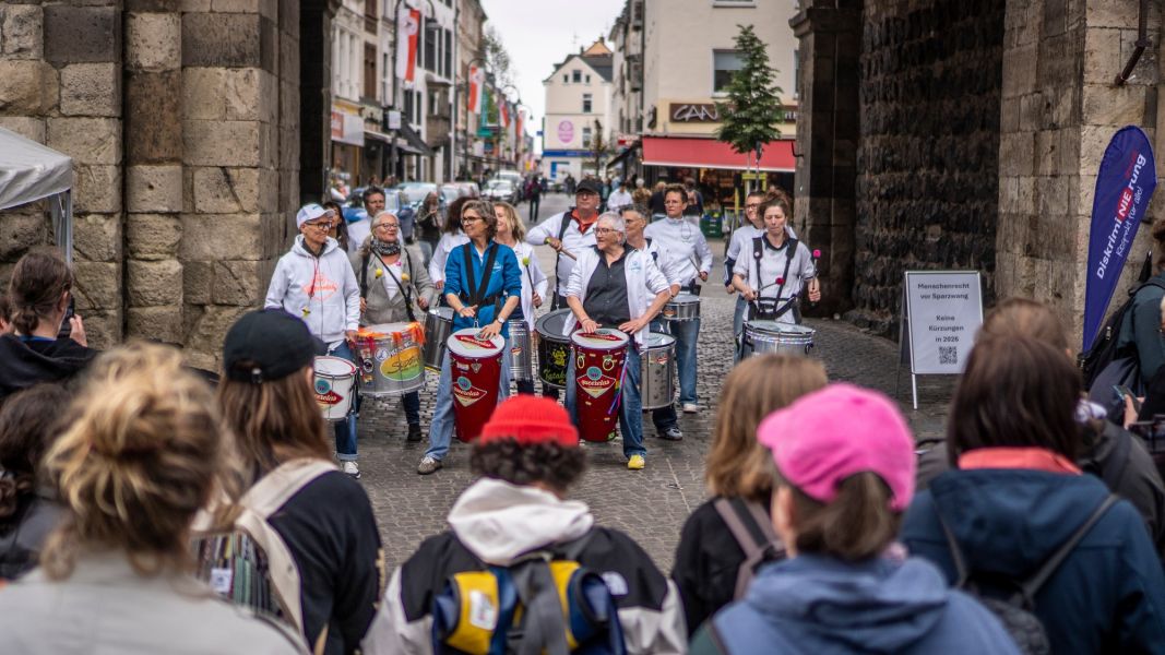 Eine Gruppe von Frauen trommelt auf großen Trommeln. Im Vordergrund (unscharf) stehen einige Zuschauer*innen, die der Gruppe zugewandt sind. 