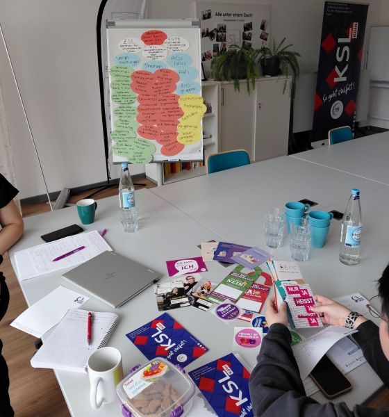 Das Foto zeigt von oben einen Konferenztisch, auf dem zahlreiche Flyer und Broschüren des KSL.Köln und Kooperationspartner*innen präsentiert werden. Auch Gläser, Tassen, ein Laptop und Schreibutensilien liegen dort. Zwei Personen sind zum Teil zu sehen; eine hält Flyer in der Hand. Im Hintergrund steht ein Flipchart mit vielen Moderationskarten darauf. 