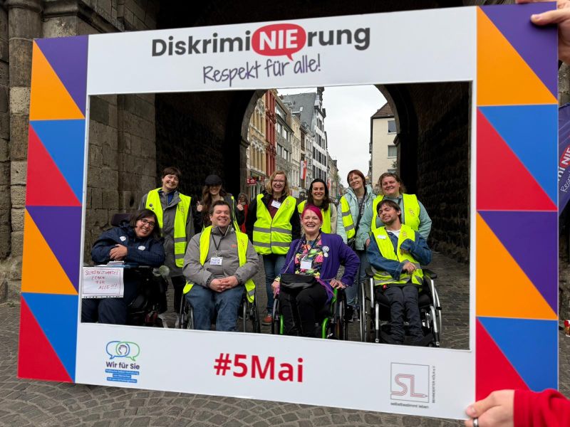 Durch einen großen, tragbaren Bilderrahmen, auf dem der Veranstaltungstitel, die Logos der Veranstaltenden und "#5Mai" steht, wird vor eine Gruppe von zehn Personen aus dem Veranstaltungsteam in gelben Warnwesten fotografiert.