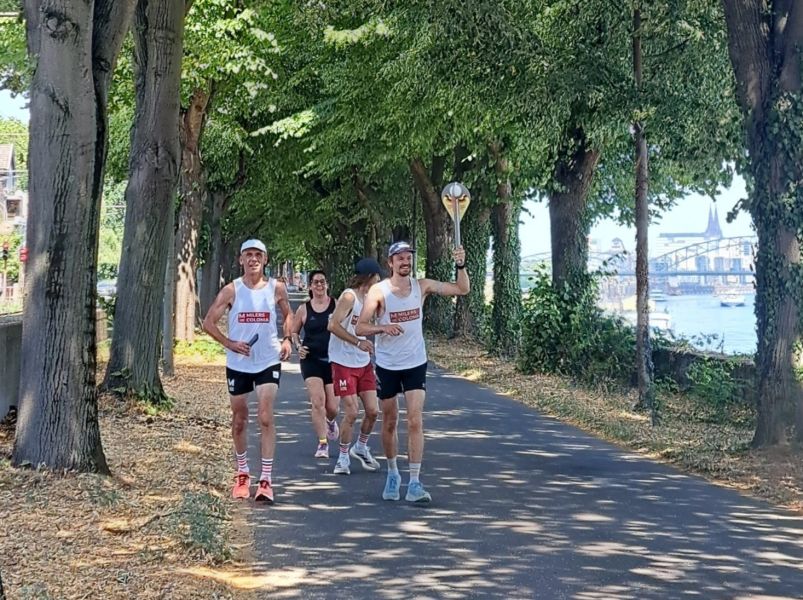 Vier Läufer*innen laufen durch eine Baumallee am Rhein. Ein Läufer hält den Staffelstab nach oben.