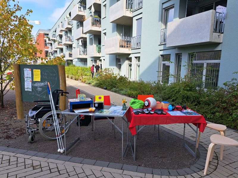 Im Innenhof des Vereins stehen zwei Tische, auf dem unter anderem Brillen, Ohrenschutz und ein Fußball liegen, werden von weitem gezeigt. Daneben steht ein Rollstuhl, an dem drei Langstöcke angelehnt sind.