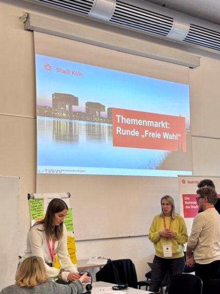 Fünf Personen stehen bzw. sitzen in einem Seminarraum um einen Tisch herum. Hinter ihnen befindet sich eine beschriftete Flipchart-Wand. An die Wand hinter ihnen wird eine Präsentation projiziert, auf der ein Bild von Köln sowie das Logo der Stadt Köln zu sehen sind. In einem Textfeld steht: "Themenmarkt: Runde "Freie Wahl".