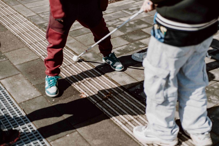 Zwei Jungen erkunden das Blindenleitsystem der Schule mit dem Langstock und erfahren so direkt, wie Menschen mit Sehbehinderungen Wege finden und welche Hindernisse im Alltag lauern können.