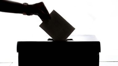 Es ist die Silhouette einer Hand zu sehen, die einen Stimmzettel in eine Wahlurne wirft.