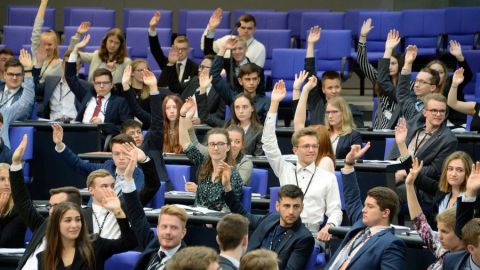 Viele Personen sitzen in einem Plenum und viele davon heben zur Abstimmung einen Arm.