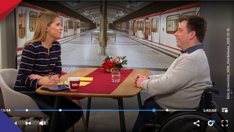 Wolfgang Thiems und Moderatorin Miriam Lange sitzen an einem Tisch und unterhalten sich über barrierefreies Reisen. Im Hintergrund ist ein Bahnsteig zu sehen, an dem auf beiden Seiten Züge stehen.