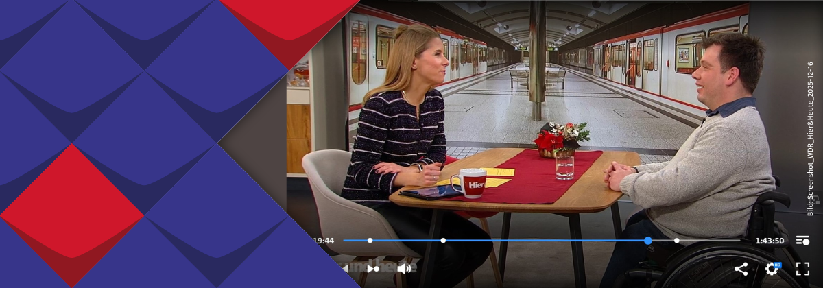 Wolfgang Thiems und Moderatorin Miriam Lange sitzen an einem Tisch und unterhalten sich über barrierefreies Reisen. Im Hintergrund ist ein Bahnsteig zu sehen, an dem auf beiden Seiten Züge stehen.