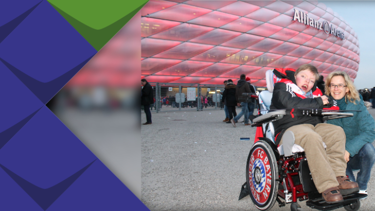 Vor der Allianz-Arena in München steht ein Rollifahrer, der sich offensichtlich freut, daneben hockt seine Mutter.