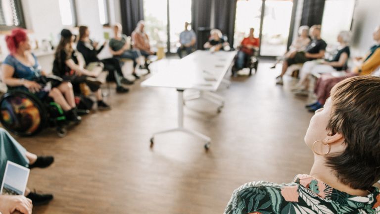 Nahaufnahme von Lena Ingenhaag, die scharf im rechten Vordergrund des Bildes steht. Im Hintergrund sind die übrigen Teilnehmenden des EUTB-Treffens als unscharfer Stuhlkreis zu erkennen.