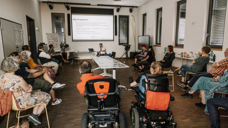 Foto eines EUTB-Netzwerktreffens in Nordrhein-Westfalen mit insgesamt 15 Teilnehmenden. Die Gruppe sitzt als offener Halbkreis nach vorne zur Präsentationsfläche ausgerichtet. Im vorderen Bereich sitzen zwei EUTB-Berater*innen in elektrischen Rollstühlen. Die übrigen dreizehn Personen nehmen die Plätze rechts und links im Halbkreis ein. Alle blicken gemeinsam auf eine digitale Präsentation von Wolfgang Thiems und dem KSL-Köln. In der Mitte des Raumes stehen zwei Tische, auf denen mehrere Fotos liegen.